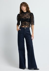 Top corto in pizzo nero con maniche a sbuffo abbinato a pantaloni larghi in denim scuro a vita alta. L'outfit presenta una chiusura a bottone e una leggera texture.