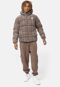 Chaqueta acolchada marrón con intrincados patrones blancos y capucha. Combinada con pantalones cargo marrones y zapatillas blancas, con una textura suave.