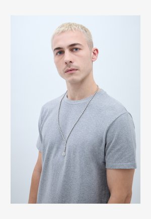 Jaunuolis trumpais blondinų plaukais, dėvintis pilką marškinėlį ir sidabrinę karoliukų grandinėlę su apvaliu pakabuku, priešais paprastą šviesų foną.