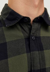 Gros plan d'une personne portant une chemise à carreaux noire et vert foncé, avec un col et deux boutons visibles attachés.