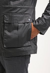 Czarna nylonowa kurtka z plisowaną kieszenią z przodu zapinaną na zatrzaski i gładką fakturą, noszona na ciemnym dżinsie.