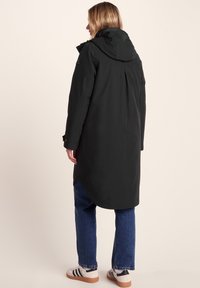 Woman wearing a black hooded long coat, blue jeans, and white sneakers with black stripes facing away against a plain background.