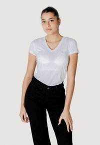 T-shirt bianco attillato con scollatura a V e un logo sottile di colore verde. Abbinato a pantaloni neri con vita alta. Materiale in misto cotone, texture liscia.