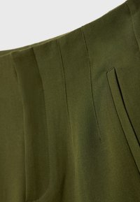 Gros plan sur un pantalon tailleur vert olive montrant la ceinture, les coutures verticales et une poche avant en biais sur tissu lisse.