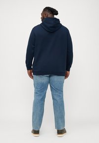 Marineblauwe hoodie met een voorzak en een capuchon met koord, gecombineerd met lichtblauwe jeans. De stof lijkt zacht te zijn met een ontspannen pasvorm.