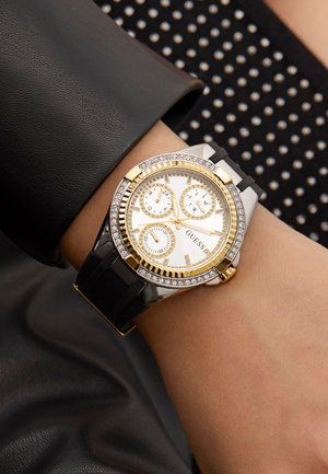 Montre dorée et argentée avec un cadran blanc, trois sous-cadrans, des accents en cristal sur la lunette et un bracelet noir, portée sur un poignet.