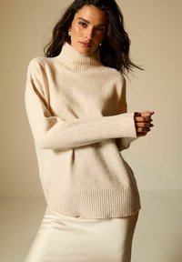 Beżowy wełniany sweter z golfem, z ryflowanymi mankietami i dołem, o luźnym kroju i miękkiej fakturze. Połączony z gładką, jasnokolorową spódnicą.