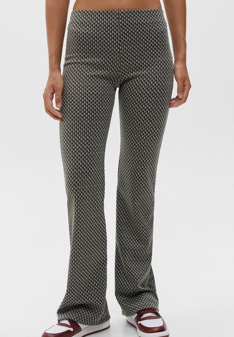 Femme portant un pantalon taille haute évasé avec un petit motif géométrique et des baskets blanches et bordeaux, debout devant un fond uni.