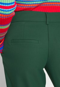 Gröna skräddarsydda byxor gjorda av texturerat tyg, med en platt bakficka. Delvis synlig är en flerfärgad randig tröja ovanpå.