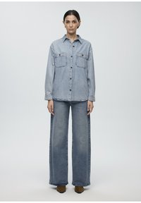 Light blue denim shirt with two chest pockets, button-down front, relaxed fit, paired with wide-leg denim jeans and brown ankle boots.