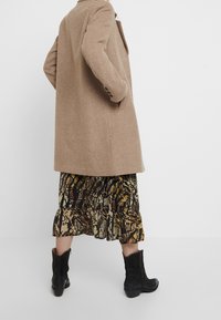 Persona che indossa un cappotto di lana cammello, una gonna midi con stampa serpente e stivali neri alla metà del polpaccio, in piedi davanti a uno sfondo bianco uniforme.