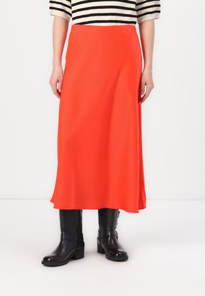 Osoba ubrana w jaskrawo pomarańczową spódnicę o długości do połowy łydki, czarne kozaki za kolano oraz czarno-białą prążkowaną bluzkę, stojąca na tle jednolitego białego tła.