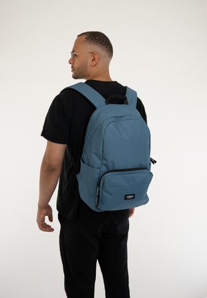 Rucsac albastru din material textil, cu bretele reglabile, un buzunar frontal cu fermoar și un patch cu logo. Textură netedă și design modern, potrivit pentru utilizare casuală.