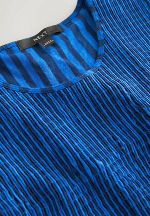 Mėlynos tekstūruotos medžiagos viršus su tamsesnėmis mėlynomis vertikaliomis juostelėmis ir apvalia iškirpte, pažymėtas etiketė „NEXT“.