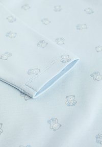 Un tissu bleu clair présente de petits motifs de ours en peluche gris et bleu. Le gros plan montre un bord replié avec une couture fine pour le détail.