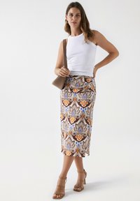 Falda midi estampada con diseño floral en marrón, naranja y azul. Top de tirantes acanalado blanco, bolso marrón y sandalias de tacón color arena. Textura suave.