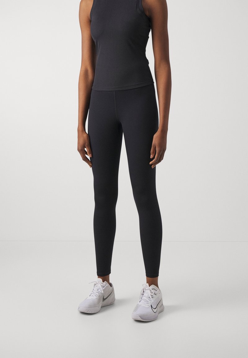 Svart sömlös träningsöverdel med hög halsringning, matchad med eleganta svarta leggings. Fötter i vita sportskor med grå accenter och snören.