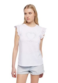 T-shirt bianco in cotone con maniche corte a volant in pizzo, caratterizzato da un design a cuore luccicante sul davanti. Abbinato a pantaloni shorts in denim chiaro.