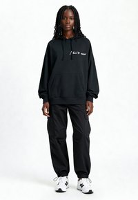 Sweat-shirt à capuche noir avec le texte "Je m'en fiche" sur le devant, associé à un pantalon cargo noir ample et des chaussures de sport blanches.