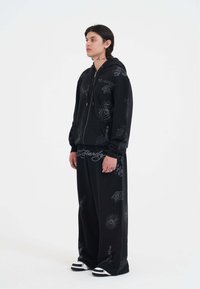 Ung person står iført en sort oversized hoodie og bukser med spindelvævs-, tiger- og tatoveringslignende grafiske tryk, hvide sneakers.