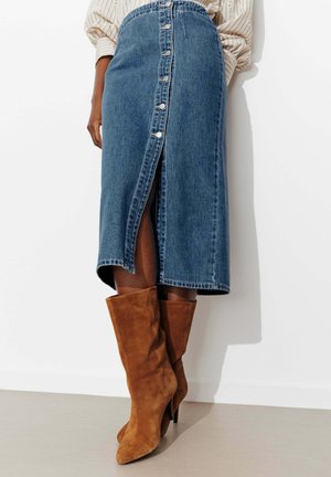 Jupe en denim bleu mi-longue avec boutons à l'avant et fente, portée avec des bottes à talons en suède marron et un chemisier beige.
