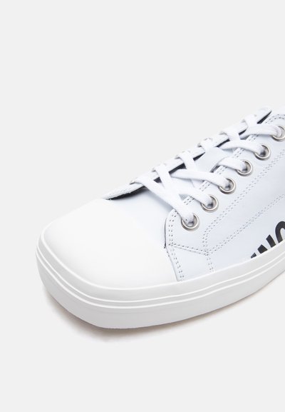 Baskets basses blanches avec des lacets blancs, des œillets en métal, des détails cousus, et un texte noir partiel sur le côté, sur un fond uni.