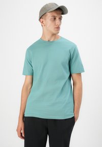 Tricou scurt cu maneca scurta de bumbac turcoaz, cu guler rotund, purtat cu pantaloni închiși la culoare, completat de o șapcă gri. Design simplu și fără imprimeuri.