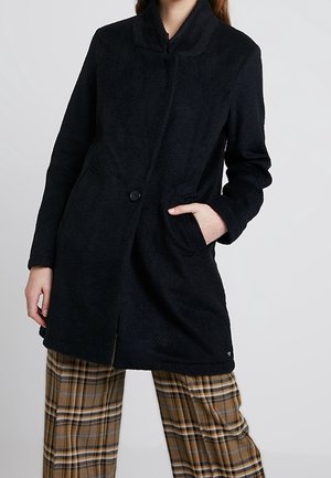 Schwarzer, langer Wollmantel mit hohem Kragen, Ein-Knopf-Verschluss und Seitentaschen. Kombiniert mit karierter Hose. Weiche Textur.