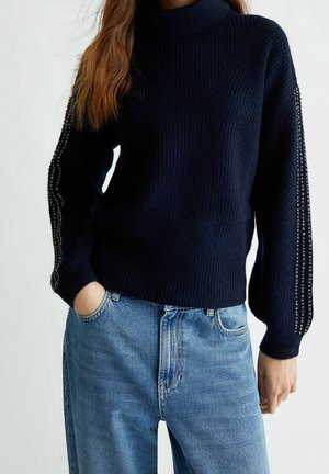 Woman wearing a navy ribbed turtleneck sweater with studded details on sleeves and light blue jeans with hand in pocket.