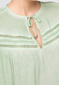 Blusa verde chiaro con scollo rotondo e chiusura con laccetto, delicati dettagli in pizzo a occhio e tessuto morbido e fluido.