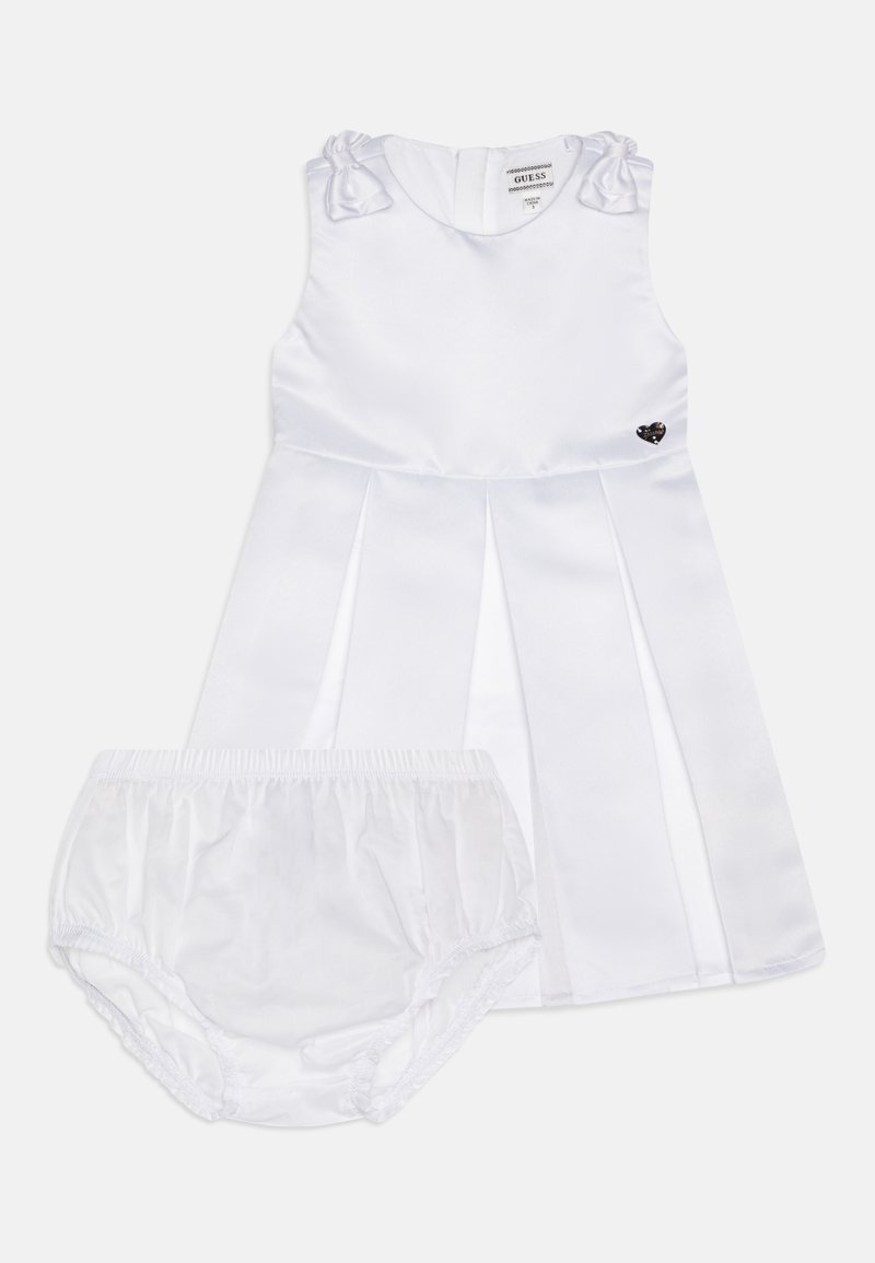 Λευκό σατέν φόρεμα με πτυχώσεις, φιογκάκια στους ώμους και μια μικρή καρδιά ως διακόσμηση. Περιλαμβάνει αντίστοιχες λευκές βερμούδες.