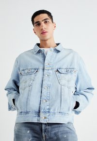 Chaqueta de mezclilla azul claro con un diseño clásico de botones, bolsillos en el pecho y un ajuste relajado con detalles de costura visibles.
