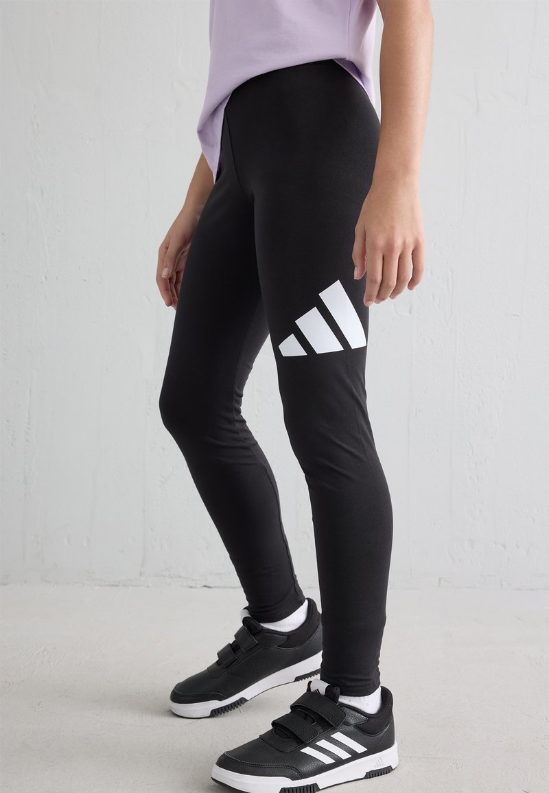 Personne portant des leggings noirs avec un logo blanc et des baskets à velcro noires avec des semelles blanches, se tenant devant un arrière-plan clair.