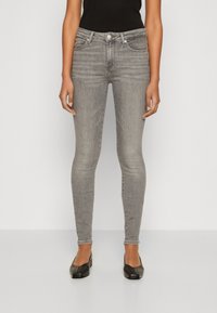 Mujer llevando jeans grises ajustados, top negro y zapatos de tacón bajo negros, de pie contra un fondo claro y simple.