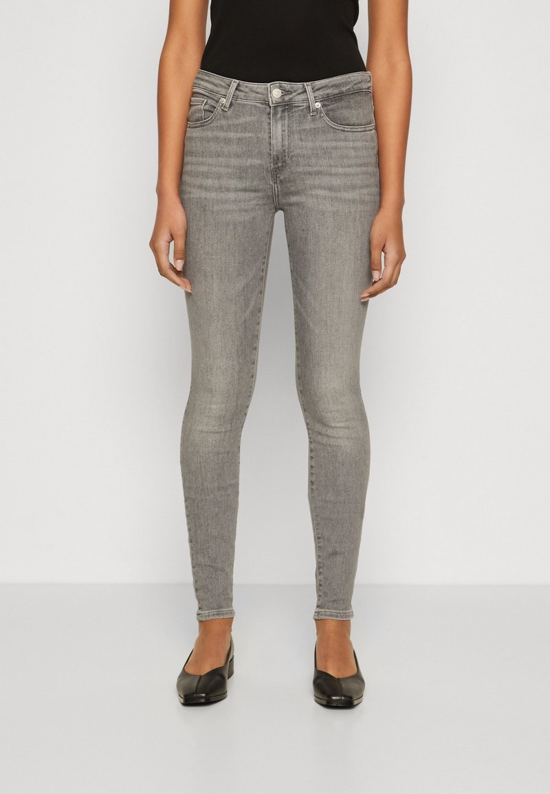 Mujer llevando jeans grises ajustados, top negro y zapatos de tacón bajo negros, de pie contra un fondo claro y simple.