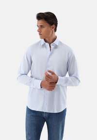 Helles blaues Langarmhemd mit Knopfleiste, aus Baumwollstoff, mit umgeschlagenem Kragen, vorderer Knopfleiste, abgerundetem Saum, getragen mit Jeans.