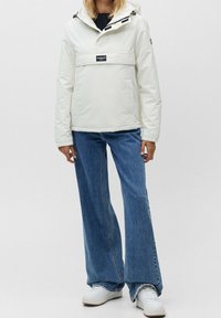 Vit huva jacka med en framficka, svart logotyp. Kombinerad med utsvängda blå jeans och vita sneakers. Mjuk, slät textur.