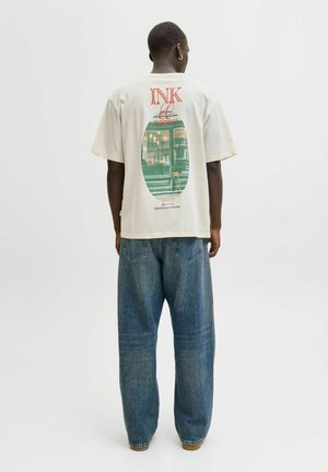 Persona che indossa una T-shirt crema con una grafica di una finestra di caffetteria e il testo "INK & espresso" sul retro, abbinata a jeans blu.