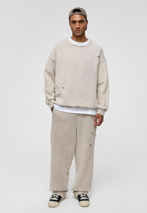 Beige oversized sweatshirt en bijpassende broek met versleten details en een textuurafwerking; gedragen met een wit T-shirt en zwarte sneakers.