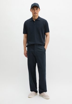 Man in zwart poloshirt, zwarte broek, zwarte pet en lichtbeige sneakers, staand met één hand in de zak.