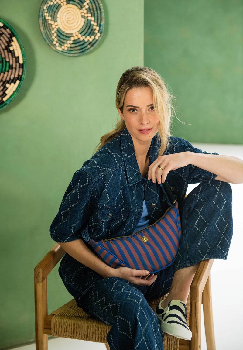 Denim outfit with diamond patterns, blue and white striped bag, wooden chair, and textured wall in green tones.