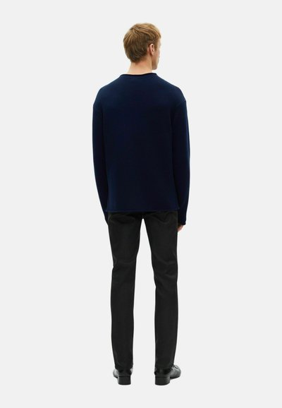 Marineblauer Strickpullover mit Rundhalsausschnitt, lässiger Schnitt, strukturierter Stoff, kombiniert mit schwarzen Hosen. Ansicht von hinten, Person steht mit den Händen an den Seiten.