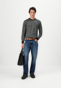 Črna pulover s ponavljajočim se vzorcem logotipa, v kombinaciji z modrimi jeans hlačami. Model drži črno torbico, oblečen v črne čevlje z teksturirano površino.