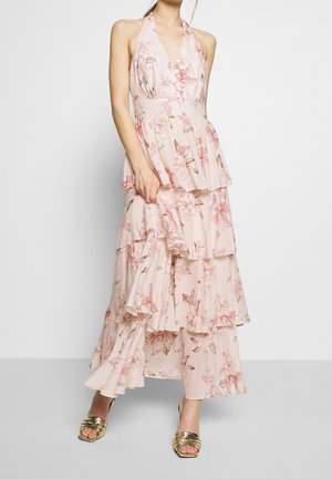 Lange halterjurk met bloemenprint in lichtroze, voorzien van gelaagde ruches, een getailleerde taille en delicate bloemenpatronen. Draagt hierbij gouden sandalen met hak.