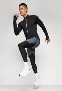 Haut de sport noir à manches longues avec une fermeture éclair vert fluo, associé à un short gris et des leggings noirs. Chaussures de course blanches et chaussettes visibles.