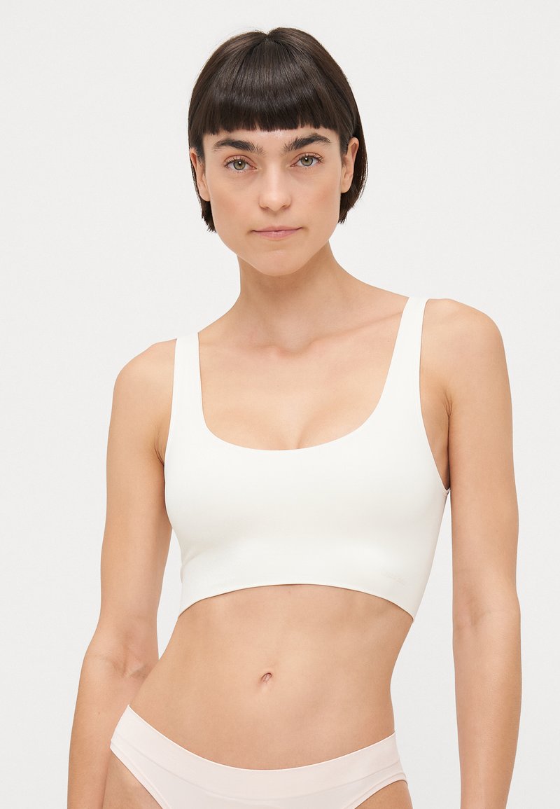 Crop top bianco senza cuciture con spalline larghe, scollatura quadrata e una texture liscia e elastica. Indossato con biancheria intima rosa chiaro.