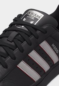 Μαύρο παπούτσι Adidas με λευκές και κόκκινες ρίγες, μαύρα κορδόνια και λογότυπο Messi στο πλάι κοντά στα κορδόνια.