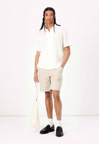 Vit kortärmad skjorta med vertikal textur, beige randiga shorts, vita strumpor, svarta loafers och en krämfärgad tote-väska.