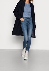 Abrigo navy sobre un suéter de punto ligero azul, jeans skinny azul oscuro con dobladillos deshilachados y botas tobilleras blancas. Atuendo simple, moderno y casual.