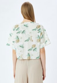 Korte blouse met cropped mouwen in lichte stof, voorzien van een groen en beige botanisch patroon met bladeren en schelpen op een witte achtergrond.